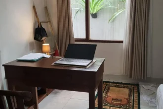 A furnished studio room rented monthly in Southeast Asia. The image shows how the tenant has a table with laptop on it and chair faced looking outside a window. There is a muslim prayer mat, which is fitting due to the location being Indonesia. The floor is tile, which is very common in Southeast Asia.