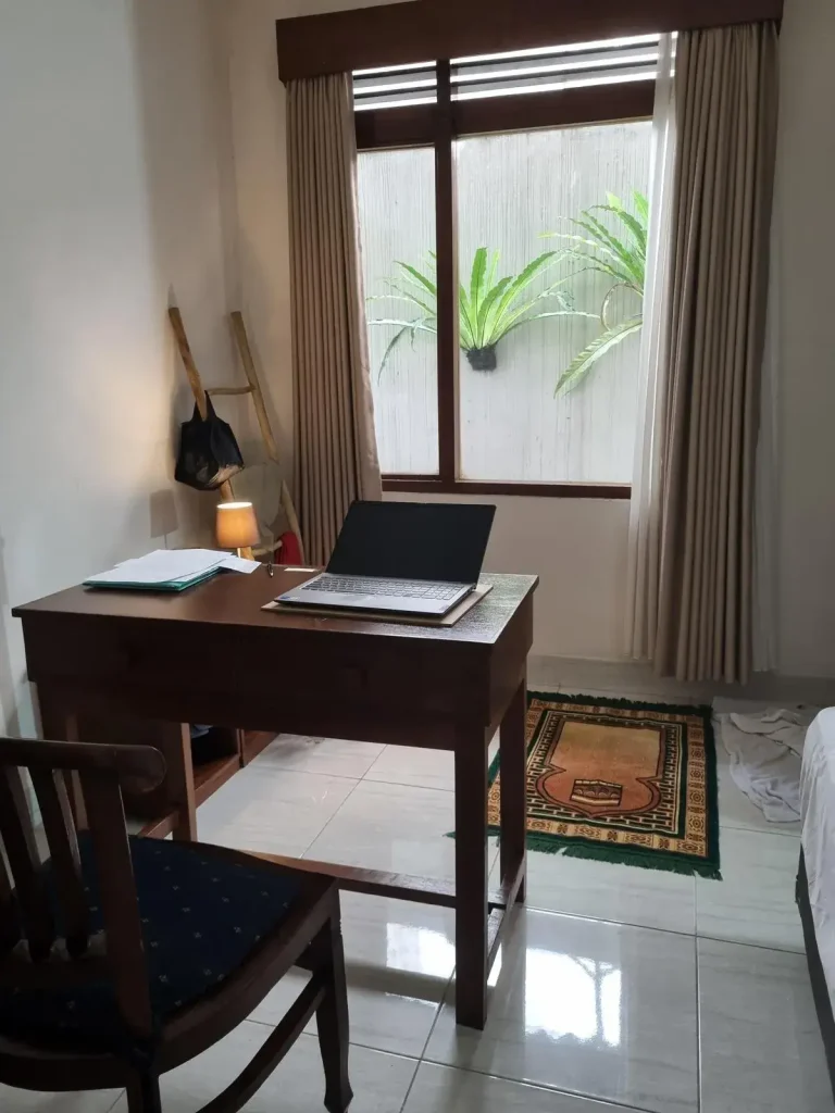 A furnished studio room rented monthly in Southeast Asia. The image shows how the tenant has a table with laptop on it and chair faced looking outside a window. There is a muslim prayer mat, which is fitting due to the location being Indonesia. The floor is tile, which is very common in Southeast Asia.
