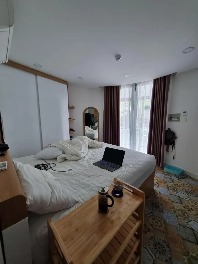 A furnished studio room rented monthly in Southeast Asia. The space is tight, but it is very well laid out and cozy. The image shows a bed with sheets, a small table pulled next to the bed with coffee on it, a split-duct airconditioning unit above the bed, a large wardrobe along one wall with extra shelving at it's side, and a full-sized mirror. Next is the front door, which is fully see-through glass, but has two sets of curtains - one that lets light in with some visibility, and another to block all light. The floor is tile, which is very common in Southeast Asia.