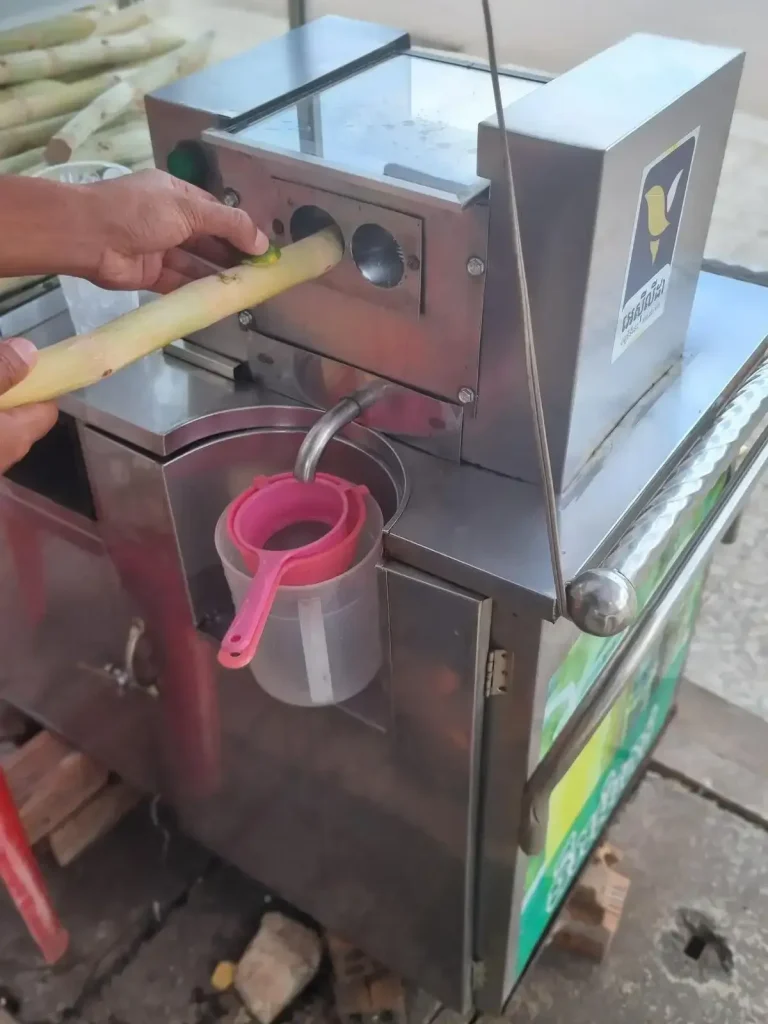 A worker using a machine to make sugarcane juice in Southeast Asia. They are actively feeding a lime and a stick of sugarcane through a machine with rollers than compress the sugarcane to squeeze the juice out of it.