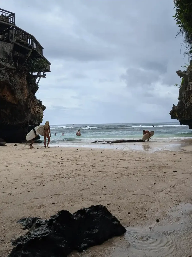 A beach within a small opening between two steep, rocky, cliffs. In the foreground, two people are walking towards the ocean water with a surfboards held at their side. In the background, people are surfing on the waves. The cliff to the left has a wooden deck on top of it, at about three stories high, for a lookout viewpoint of the ocean. This beach is located in Uluwatu, which is on the south side of Bali, Indonesia. Uluwatu is pronounced "oo-loo-wah-too".