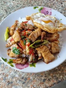 Thai stir-fried crispy pork on rice with a fried egg. The dish has Thai basil and red chillis mixed in. Crispy pork the meat as bacon, but small chunks instead of long thin slices. The meat has a thin batter on the edge, causing it to be crispy when fried.