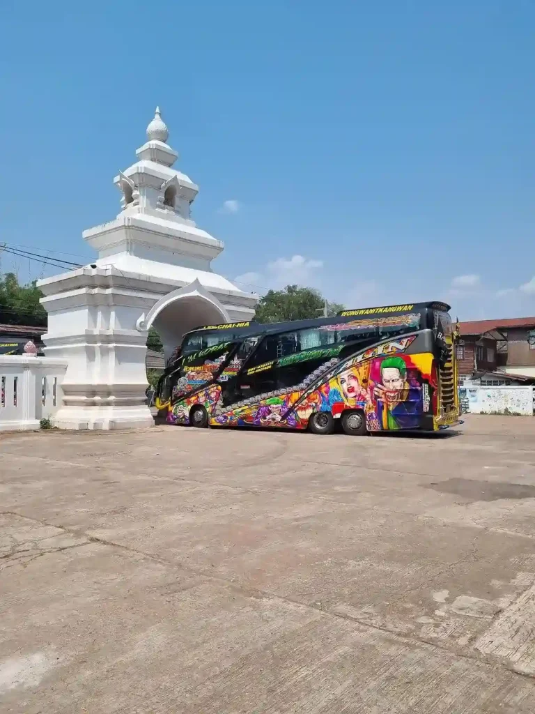 The side of a Thai (Thailand) double-decker tour bus with colorful murals on the side, including a Joker faces from Batman. The bus is driving through a temple-style entrance, and it barely fits through.