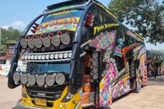 Front and side of a Thai (Thailand) double-decker tour bus with colorful murals on the side. The front has 2 horizontal rows of 6 subwoofer speakers each, a horizontal row of 13 spot lights, and a mural on the upper deck's front window.