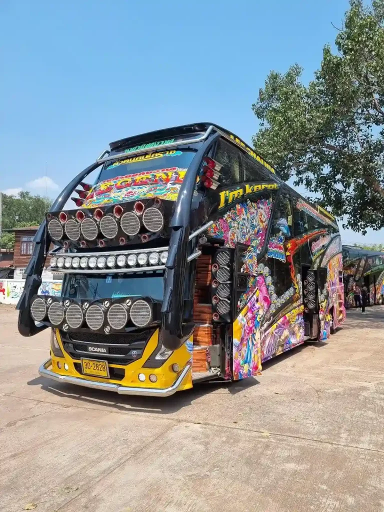 Front and side of a Thai (Thailand) double-decker tour bus with colorful murals on the side. The front has 2 horizontal rows of 6 subwoofer speakers each, a horizontal row of 13 spot lights, and a mural on the upper deck's front window.