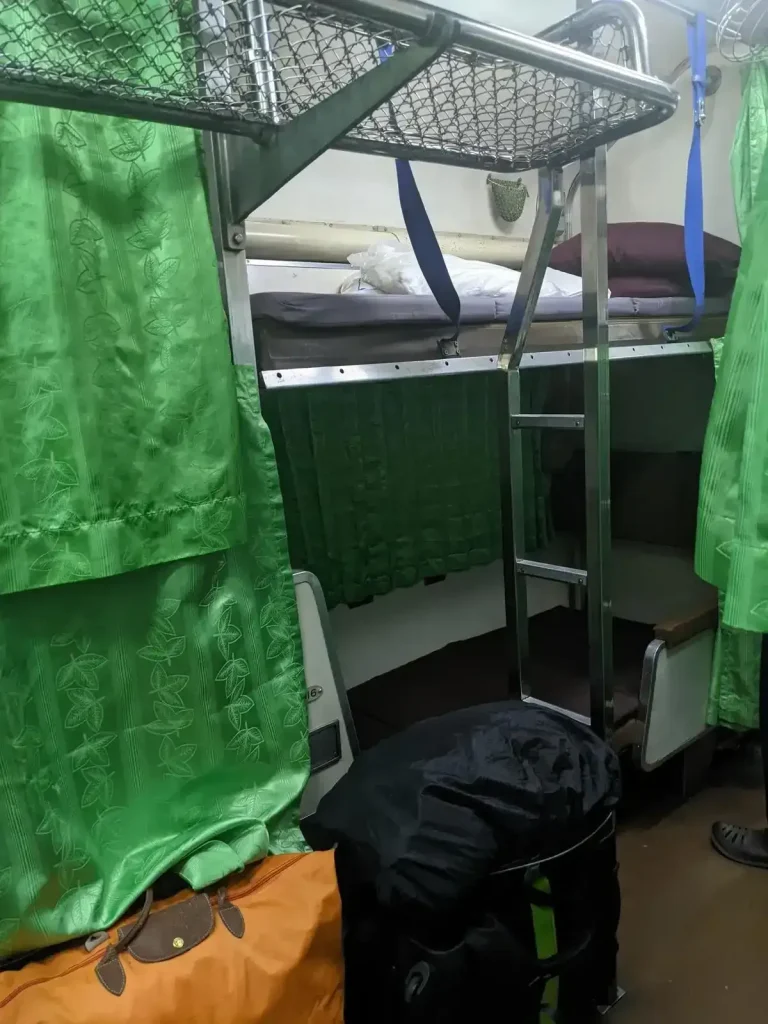 Night time bench seating that is converted to bunk beds on the inside of the Thai (Thailand) train sleeper cabin. Curtains are partially closed. Each bunk bed is for 1 adult. There is one bunk bed at the bottom, and another above with a ladder to climb up into it. There are curtains for the windows and to enclose each bunk bed, while the lighting in the aisle remains on at night. For each grouping of bench seats, there is a luggage rack to the side, and a small ladder to climb up into the upper bunk bed. The passenger has a bag that is locked and tethered to the storage rack to prevent theft.