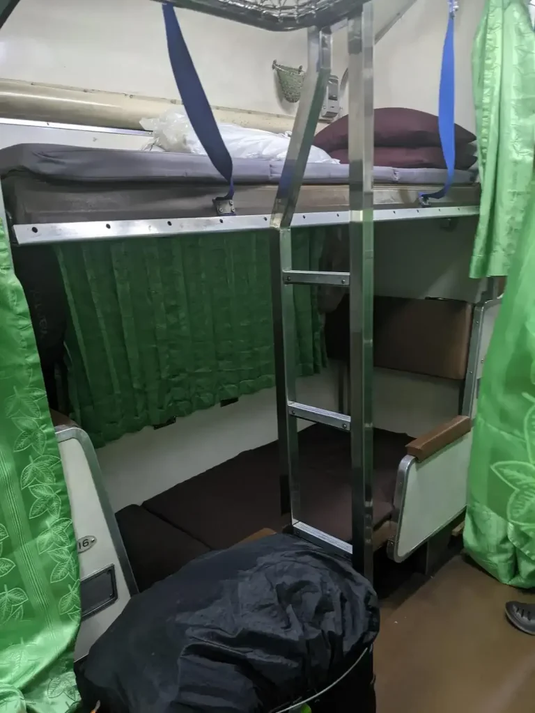 Night time bench seating that is converted to bunk beds on the inside of the Thai (Thailand) train sleeper cabin. Each bunk bed is for 1 adult. There is one bunk bed at the bottom, and another above with a ladder to climb up into it. There are curtains for the windows and to enclose each bunk bed, while the lighting in the aisle remains on at night. For each grouping of bench seats, there is a luggage rack to the side, and a small ladder to climb up into the upper bunk bed. The passenger has a bag that is locked and tethered to the storage rack to prevent theft.