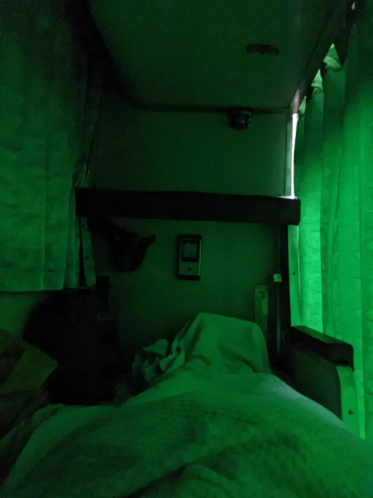 Inside the bunk beds on the inside of the Thai (Thailand) train sleeper cabin at night time. Curtains are fully closed and the bunk is dark, with lighting slightly bleeding through the curtains that enclose the singe bunk bed. Each bunk bed is for 1 adult. The passenger decided to keep their baggage inside the bunk bed with them at time to hold onto their belongings, while another bag is locked and tethered to the storage rack to prevent theft.