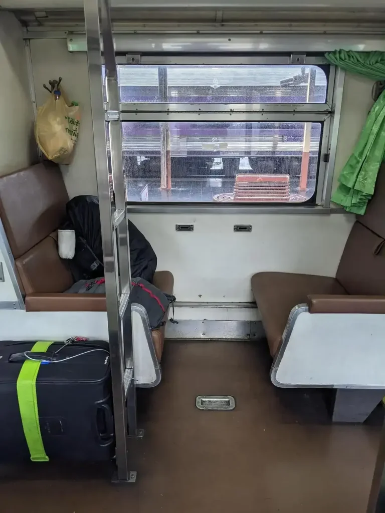 Day time bench seating on the inside of the Thai (Thailand) train sleeper cabin. During the day, each bench seat can seat 1 adults. The bench seating is in groups of 2 bench seats for 2 adults total, one bench seat facing forward plus another facing rearward. There is a good amount of leg space in between. The layout arrangement is bench seating on the left-hand side, then a middle row ailse, and another set of bench seats on the right-hand side. Windows slide open for a cool breeze. For each grouping of bench seats, there is a luggage rack to the side, and a small ladder for when the seating gets converted to bunk beds at night time. The passenger decided to keep some baggage on the bench seat with them to closely hold onto their belongings, while another bag is locked and tethered to the storage rack to prevent theft.