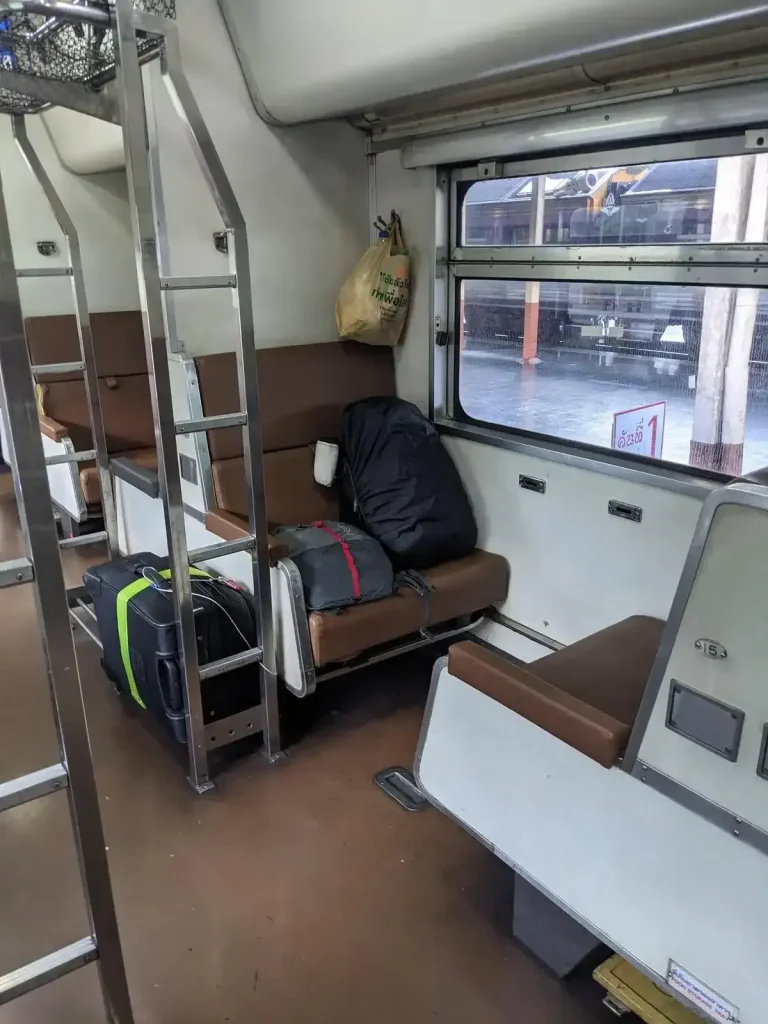 Day time bench seating on the inside of the Thai (Thailand) train sleeper cabin. During the day, each bench seat can seat 1 adults. The bench seating is in groups of 2 bench seats for 2 adults total, one bench seat facing forward plus another facing rearward. There is a good amount of leg space in between. The layout arrangement is bench seating on the left-hand side, then a middle row ailse, and another set of bench seats on the right-hand side. Windows slide open for a cool breeze. For each grouping of bench seats, there is a luggage rack to the side, and a small ladder for when the seating gets converted to bunk beds at night time. The passenger decided to keep some baggage on the bench seat with them to closely hold onto their belongings, while another bag is locked and tethered to the storage rack to prevent theft.
