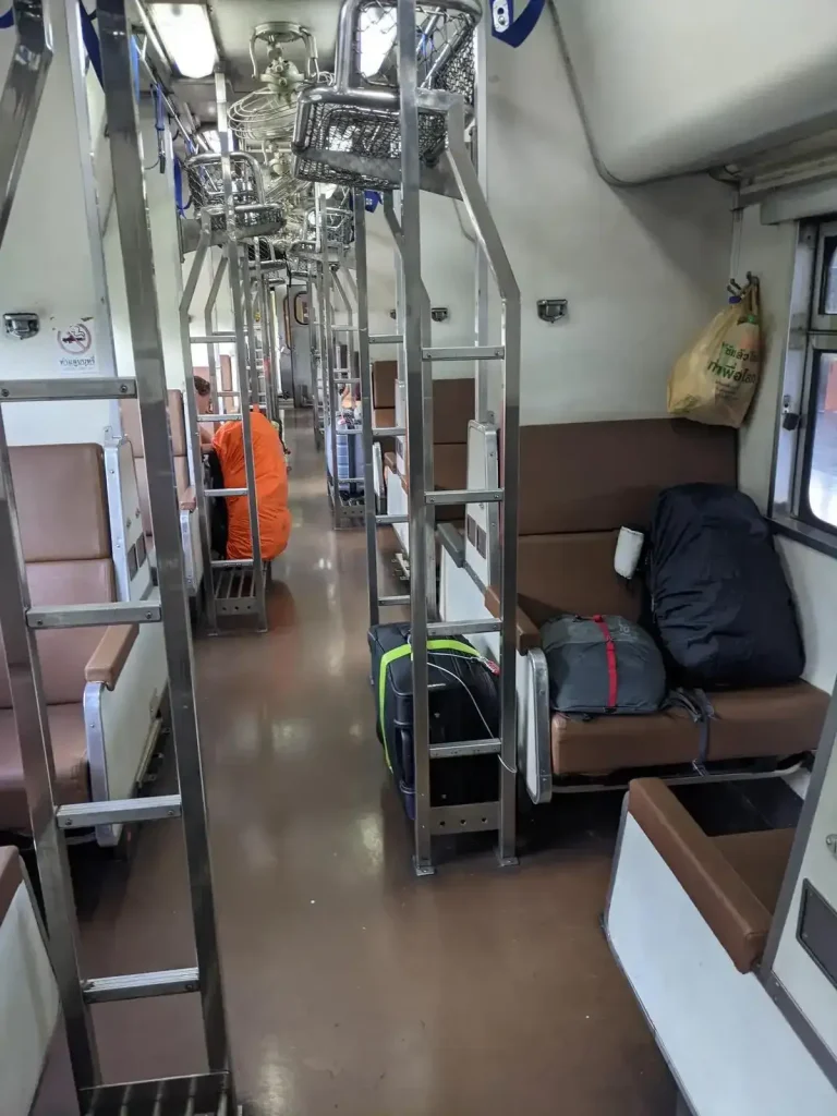 Day time bench seating on the inside of the Thai (Thailand) train sleeper cabin. During the day, each bench seat can seat 1 adults. The bench seating is in groups of 2 bench seats for 2 adults total, one bench seat facing forward plus another facing rearward. There is a good amount of leg space in between. The layout arrangement is bench seating on the left-hand side, then a middle row ailse, and another set of bench seats on the right-hand side. Windows slide open for a cool breeze. For each grouping of bench seats, there is a luggage rack to the side, and a small ladder for when the seating gets converted to bunk beds at night time. The passenger decided to keep some baggage on the bench seat with them to closely hold onto their belongings, while another bag is locked and tethered to the storage rack to prevent theft.