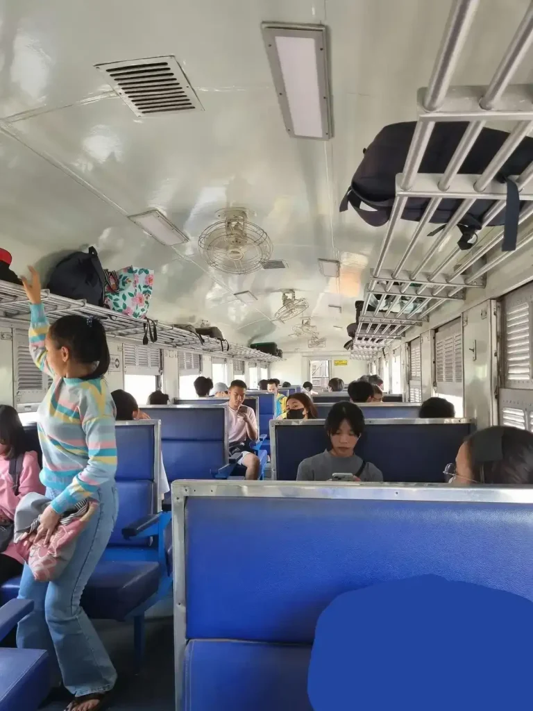 Bench seating on the inside of the Thai (Thailand) train cabin. Each bench seat can seat 2 adults. The bench seating is in groups of 2 bench seats for 4 adults total, one bench seat facing forward plus another facing rearward. There is a little bit of leg space in between. The layout arrangement is bench seating on the left-hand side, then a middle row ailse, and another set of bench seats on the right-hand side. There are several natual-air fans mounted on the ceiling. In addition, thw windows slide down for a cool breeze. There is a luggage rack mounted above all of the seats.