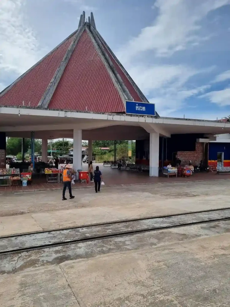 A full picture of the Kampot Campboda train stations showing the roof, station sign, passenger waiting area, food sellers with snacks on a table, and a small glimpse of the ticket office in the background. The station is open-air with no walls on the font and rear. This allows a cool breeze to flow through it on a hot day.