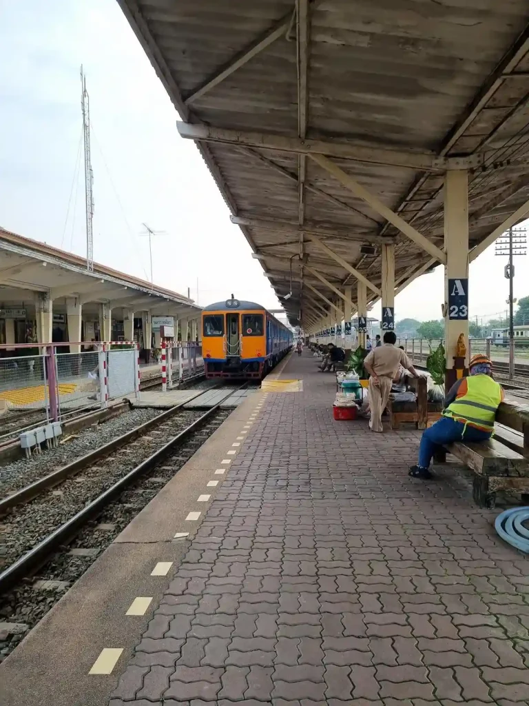 Passenger waiting area at a typical Thai (Thailand) train station. The waiting area is outside with a roof and seating underneath on a long sightly elevated concrete pad that extends the full length of the train. The rail track is to the side, along the waiting area. There are several railway staff available for assisting passengers.