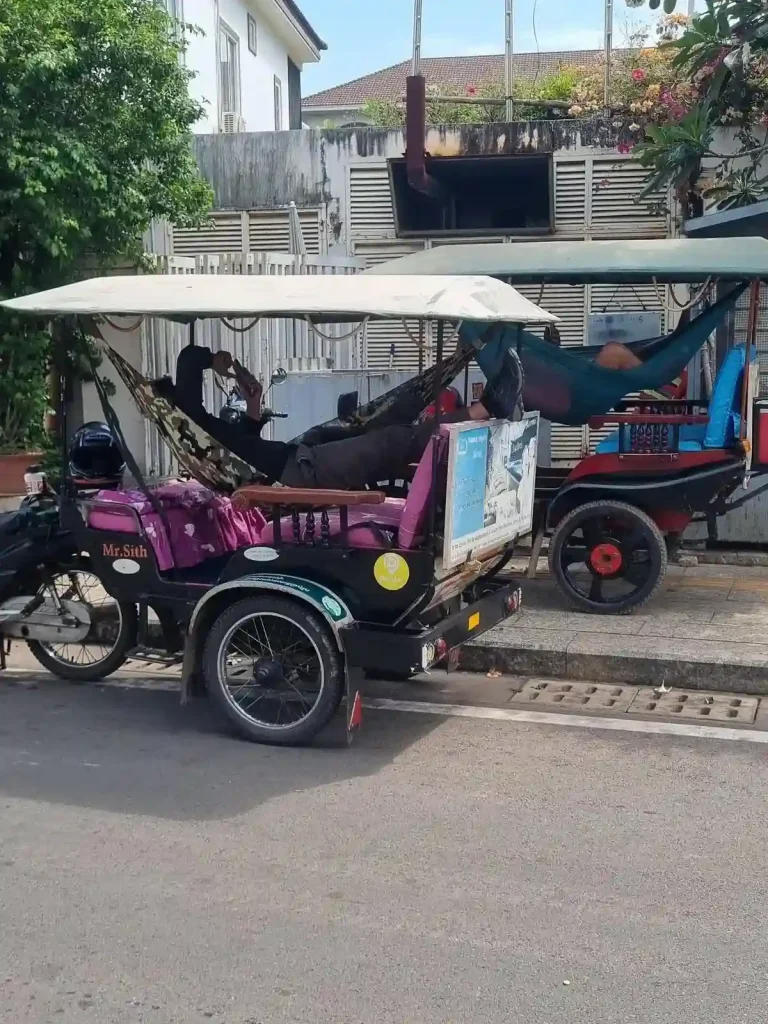Two tuk-tuks (the remorque "waggon" version) are parked next to each other. Each driver are laying in a hammock that is mounted and suspended inside the tuk-tuk passenger seating compartment. They are watching videos on their cell phones.