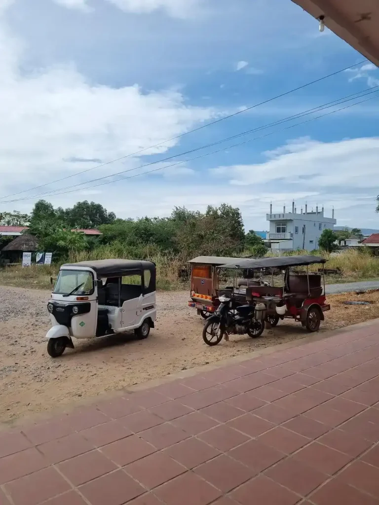 Three tuk-tuks waiting for customers outside a train station in Cambodia. There is one Indian-style three-wheel tuk-tuk, and two remorques (French word for wagon).