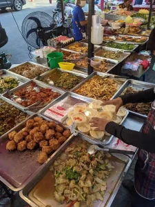 A street food stall in Bangkok, Thailand with about twenty local dishes, each on individual pans. The pans are placed against one another along a few tables, allowing customers to see everything on display and choose their next meal.