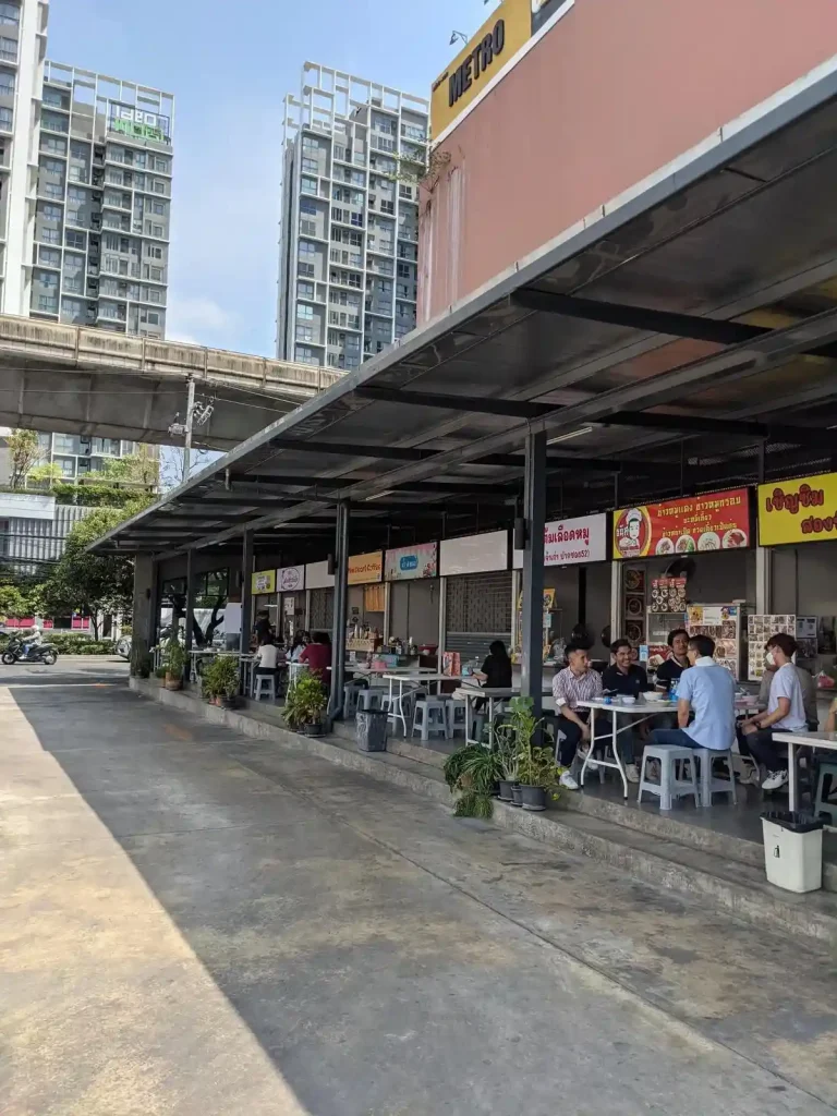 A strip of local food stalls in a clean environment in On Nut area of Bangkok, Thailand. Several office workers are eating lunch. Located off of the main street, there is a strip of food stalls that resemble outdoor storage units - each unit being a food stall - with a large overhead covering that has tables and seating underneath. The tables & seating are directly in front of the food stalls.