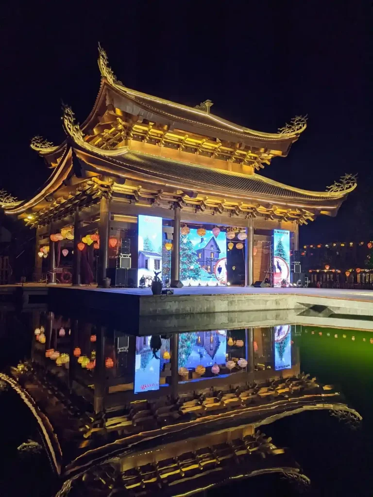 A stage during Christmas time in Vietnam. The stage is it's own building, with intricate chinese-style architectural highlights. It is built within a pond, and has a bridge connecting it to the shore.