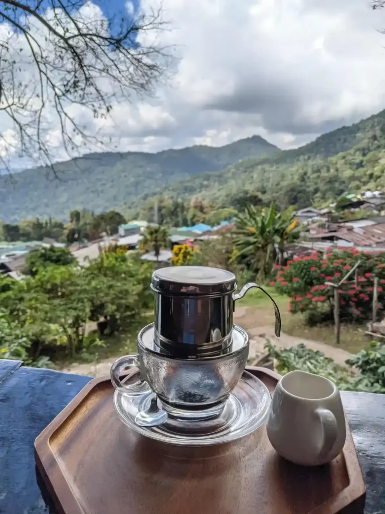 A Vietnamese Phin (pronounced "feen" as in the ending of the wording "caffeine") resting on top of a glass cup, brewing coffee, with an view of mountains in the background. This coffee filter is filled with ground coffee beans, and hot water is added inside. It uses gravity to slowly percolate (drip) hot water through the coffee grounds. At the bottom of this filter, the hot coffee drips out into a cup. This brewing method was introduced to Vietnam by the French during the colonial occupation time period.