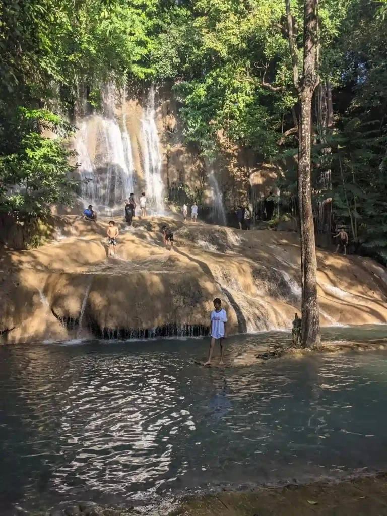 A waterfall area in Thailand surrounded by trees. The waterfall does not splace into a water pit, but onto a large rock area. The rock is limestone, and water flows over it gently. People are walking around on the rock while water flows over their feet. The water then flows off of the rock into a water pit.