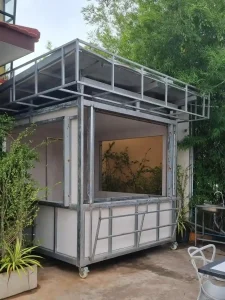 A food stall made of welded steel tubing construction in Southeast Asia. The food stall has a stell frame, and has caster wheels on all four corners. It is box shaped at a dimension of approximately six feet wide by six feet long and eight feet tall.