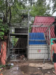 The skeleton of a small home made of welded steel tubing construction in Southeast Asia. This home resembles a tree house, in a way. It is under a tree limb, and tightly nestled between a concrete wall and a corrugated metal shed. It has two stories, with a metal ladder going to the second story. It is only about eight feet long. The section closer to the road is about eight feet wide, but because the shed is in the way, the back section of it is only about four feet wide.