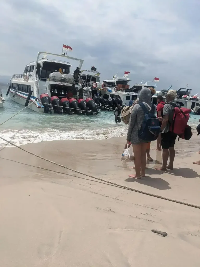 Passengers with baggage waiting on the beach to board a boat. The boat captain backs-up the boat as close to the shore as possible, but passengers and their baggage will need to walk into the water to board the boat. This boat - with 6 outboard engines - has an approximate capacity of 50 passengers, and is used to transport them from one island to another, all located within the vicinity of Bali Indonesia.