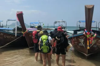 Passengers with baggage waiting in ankle-deep water close to the beach shore to board a boat. The boat captain gets front of the boat as close to the shore as possible, but passengers and their baggage will need to walk into the water to board the boat. This boat is a wooden long-tail Thai-style boat with 1 engine, and has an approximate capacity of 10 passengers. It is used to transport them from one beach to another in Ao Nang, Krabi province, Thailand.