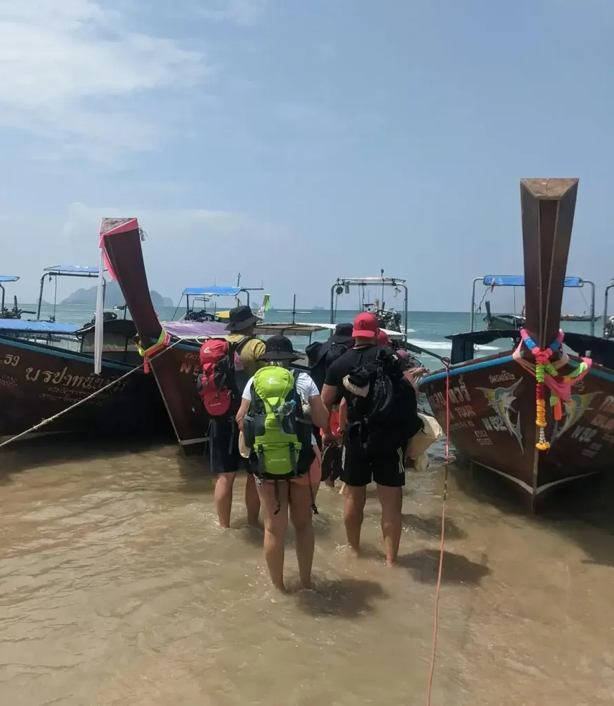 Passengers with baggage waiting in ankle-deep water close to the beach shore to board a boat. The boat captain gets front of the boat as close to the shore as possible, but passengers and their baggage will need to walk into the water to board the boat. This boat is a wooden long-tail Thai-style boat with 1 engine, and has an approximate capacity of 10 passengers. It is used to transport them from one beach to another in Ao Nang, Krabi province, Thailand.