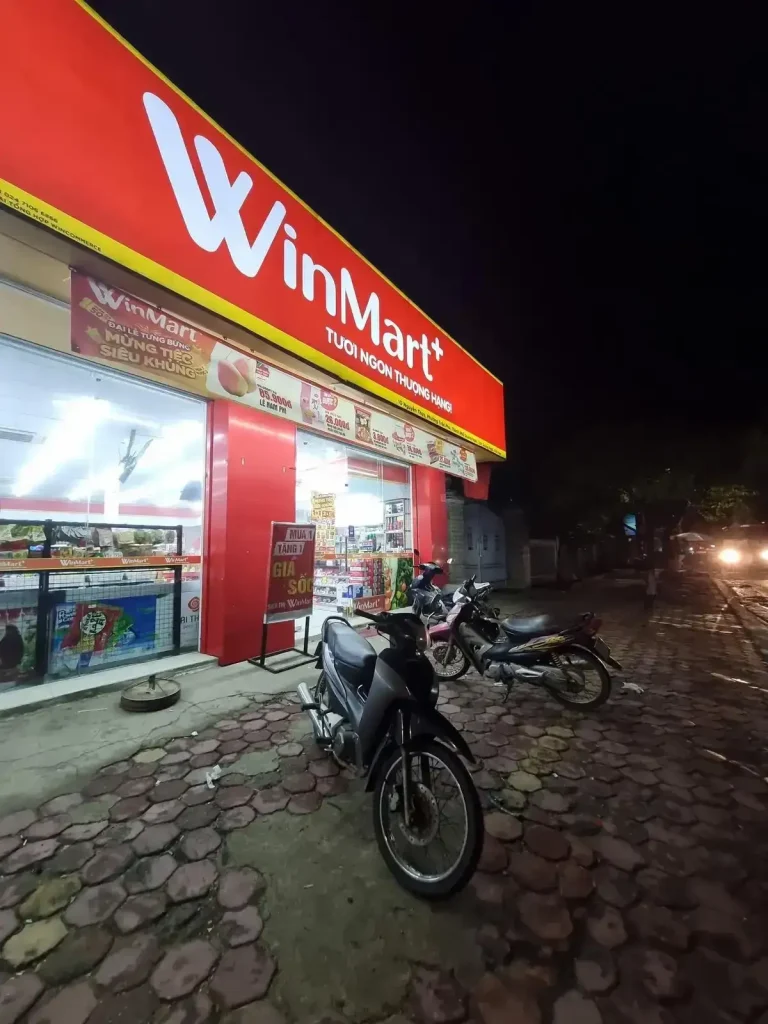 A WinMart convenience store in Vietnam with motorbike (scooter) parking on the sidewalk outside the entrance. It is night time, and the store lighting and sign is bright red.