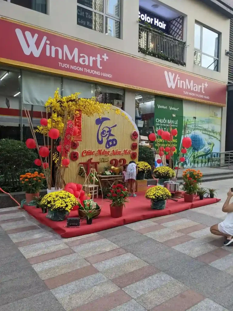 A WinMart convenience store with Tet decorations outside the store front. The decordations are a red-carpeted platform with a mix of yellow and red flowers, red Chinese lanterns, and a sign display saying "Happy New Year" in Vietnamese. It is setup for people to take pictures of themselves at. Tet is a Chinese and Vietnames lunar New Year holiday. WinMart is formerly known as VinMart, where "Vin" was short for Viet or Vietnam.
