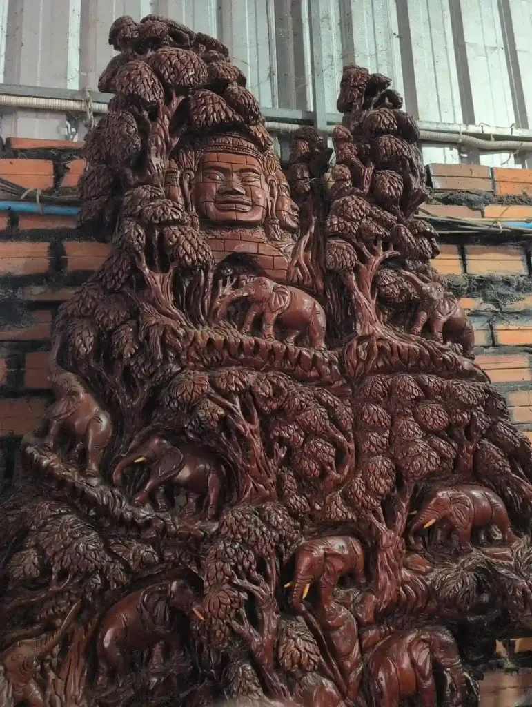 A very detailed wood carving from Cambodia of the front side of a Bayon Temple entrance gate (that has a human face on it), and several trees and elephants. The wood is a deep brown color.