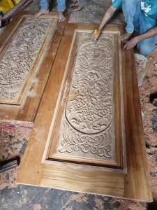 A handmade wooden door with intricate carvings in Cambodia. This image was taken inside a the shop while workers were constructing it. The workers are barefoot. One worker has a carving tool in his hand, finishing a carving.