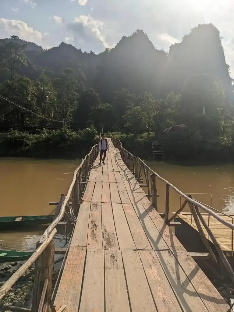 Walking on a wooden-planked bridge to cross a river in Laos. It's structure is made with a wood branches, also used to make a railing. Luscious plants and a mountainous area are waiting on the other side.