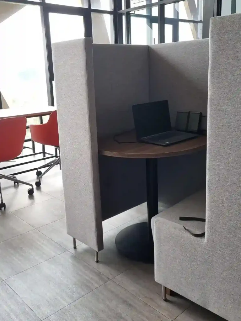A small cubicle providing private space to work at. Located in a room for office, business, and study-related activities with tables, chairs, and small cubicles. This image show a laptop on a small round desk surrounded by five feet tall walls for privacy. It also has a bench seat built into the wall structure. The edge of a conference table with roller chairs can be seen just beyond the cubicle.