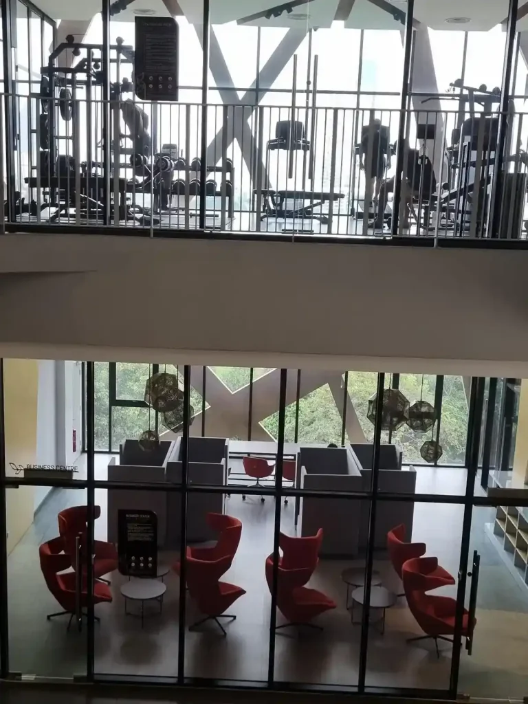An image showing rooms on two floors of amenities at an apartment complex. The top floor is a gym, and the bottom floor is a room for office, business, and study-related activities with tables (two small and one large conference table), chairs, and small cubicles (four). Both rooms have glass walls at the entrance, so it is easy to see what is inside.