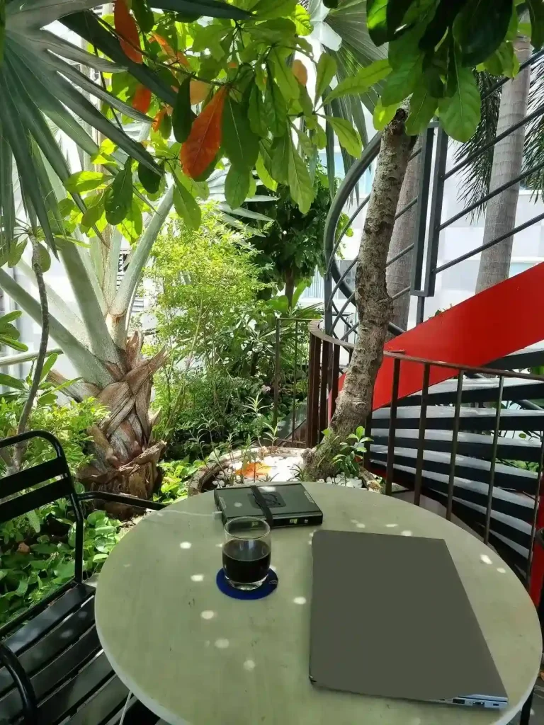 A table and chair to work at with a laptop. Plants and small trees surround the table, and provide shade. It is located on a rooftop of an apartment in Southeast Asia.