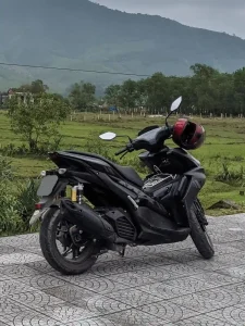 A Yamaha NMAX 155cc automatic motorbike. The motorbike is parked on a sidewalk along the road in the Vietnamese country side. There is a field and knobs (small mountains) in the background.