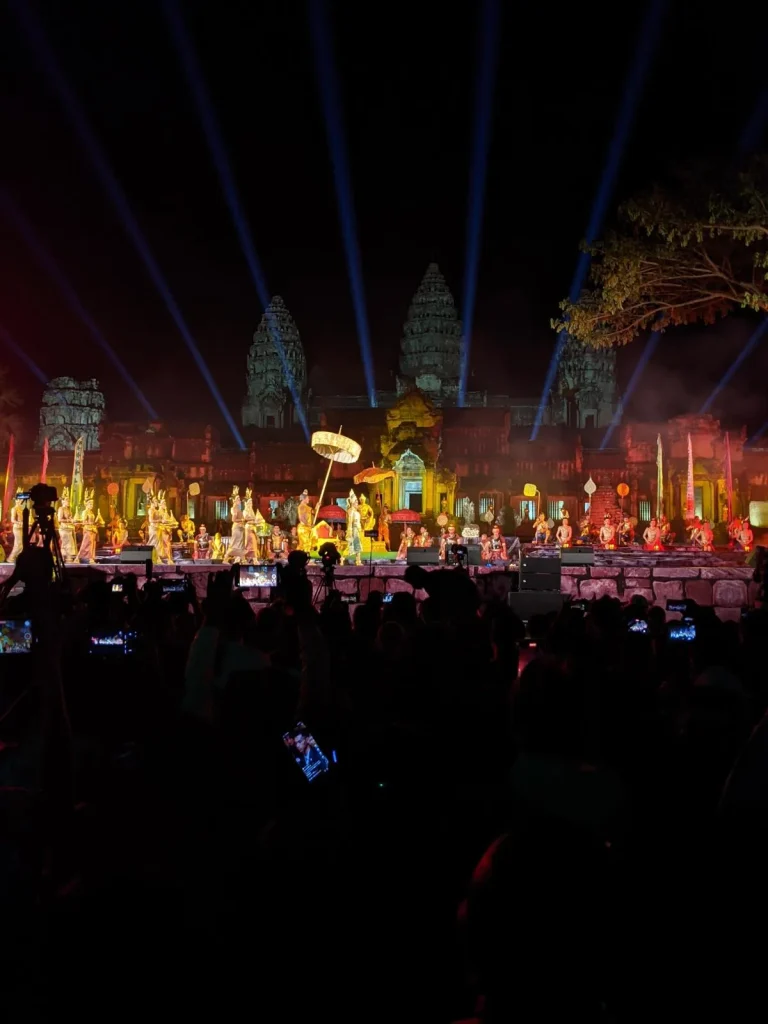 A close-up of the stage for the Angkor Thanksgiving show. On the stage, the actors are a display of the king of the Angkor empire era, with his royal parade. This show is a yearly celebration for the inclusion of Angkor Wat as a UNESCO world heritage site. It is night time, and there are lights illuminating the show stage in front of the East side of Angkor Wat. Between the photographer and stage is a crowd, many of whom have their smart phones held up to record the show and take pictures.