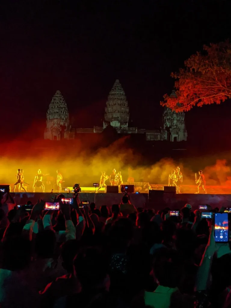A close-up of the stage for the Angkor Thanksgiving show. On the stage, there are ancient warriors battling. This show is a yearly celebration for the inclusion of Angkor Wat as a UNESCO world heritage site. It is night time, and there are lights illuminating the show stage in front of the East side of Angkor Wat. Between the photographer and stage is a crowd, many of whom have their smart phones held up to record the show and take pictures.