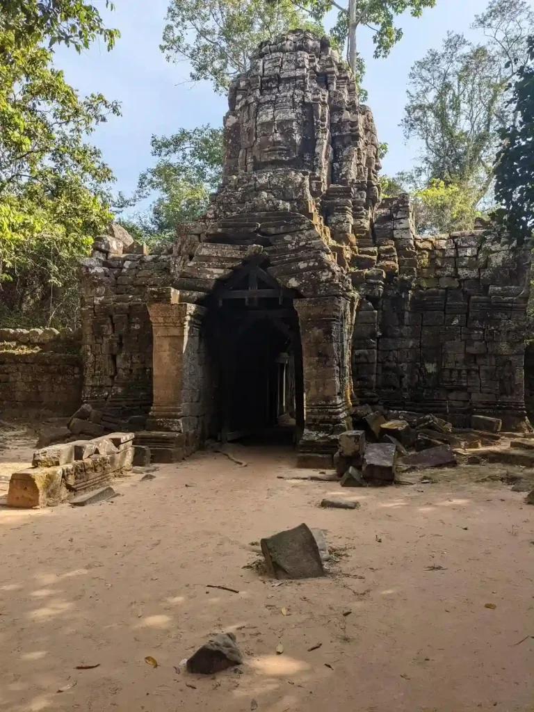 One of the four Bayon gate entrances to enter Angkor Thom. The entrance ground is all dirt, and the the gate made of stone. The top of the gate has four ancient Cambodian faces, which makes it a unique piece of archetecture. However, only one can be seen in this image. Each face is on one side, for a total of four sides - one face on each side of the entrance, one face to the left and one face to the right. This is located within Angkor Archaeological Park, which is a UNESCO World Heritage Site.
