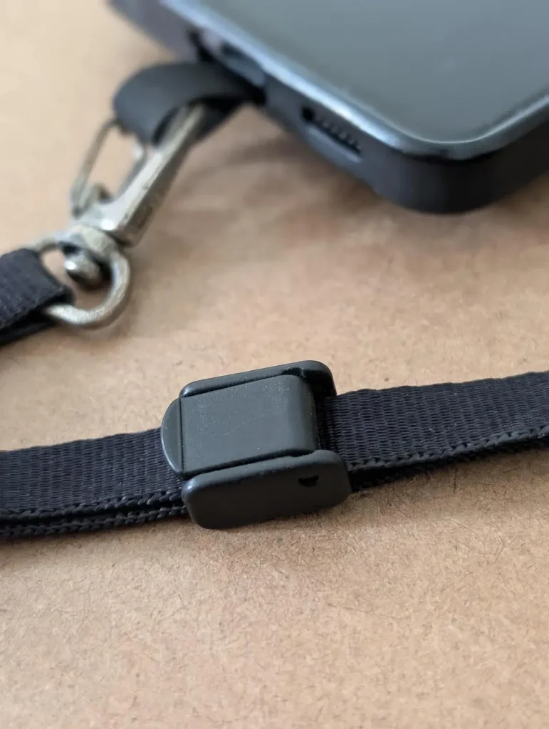 A cellphone tether tab mounted to a cellphone with a small leash attached to it. The locking clip on the leash is closed (in the lock position). This tether tab and leash combo is made by BlackRapid. The least has a quick-attach hook (similar to a rock climbing clip) on one end, and locking clip on the other end. You can slide your hand through the leash and use the locking clip to tighten it around your wrist, so that it stays on securely. Double-sided tape is provided. The tether tab is aligned with the bottom of the cellphone and meant to stick out where the charging port is. If a cellphone case is used, the tether tab will stick out through the hole for the charging port.