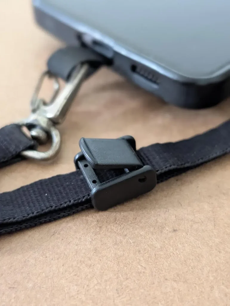 A cellphone tether tab mounted to a cellphone with a small leash attached to it. The locking clip on the leash is half closed. This tether tab and leash combo is made by BlackRapid. The least has a quick-attach hook (similar to a rock climbing clip) on one end, and locking clip on the other end. You can slide your hand through the leash and use the locking clip to tighten it around your wrist, so that it stays on securely. Double-sided tape is provided. The tether tab is aligned with the bottom of the cellphone and meant to stick out where the charging port is. If a cellphone case is used, the tether tab will stick out through the hole for the charging port.