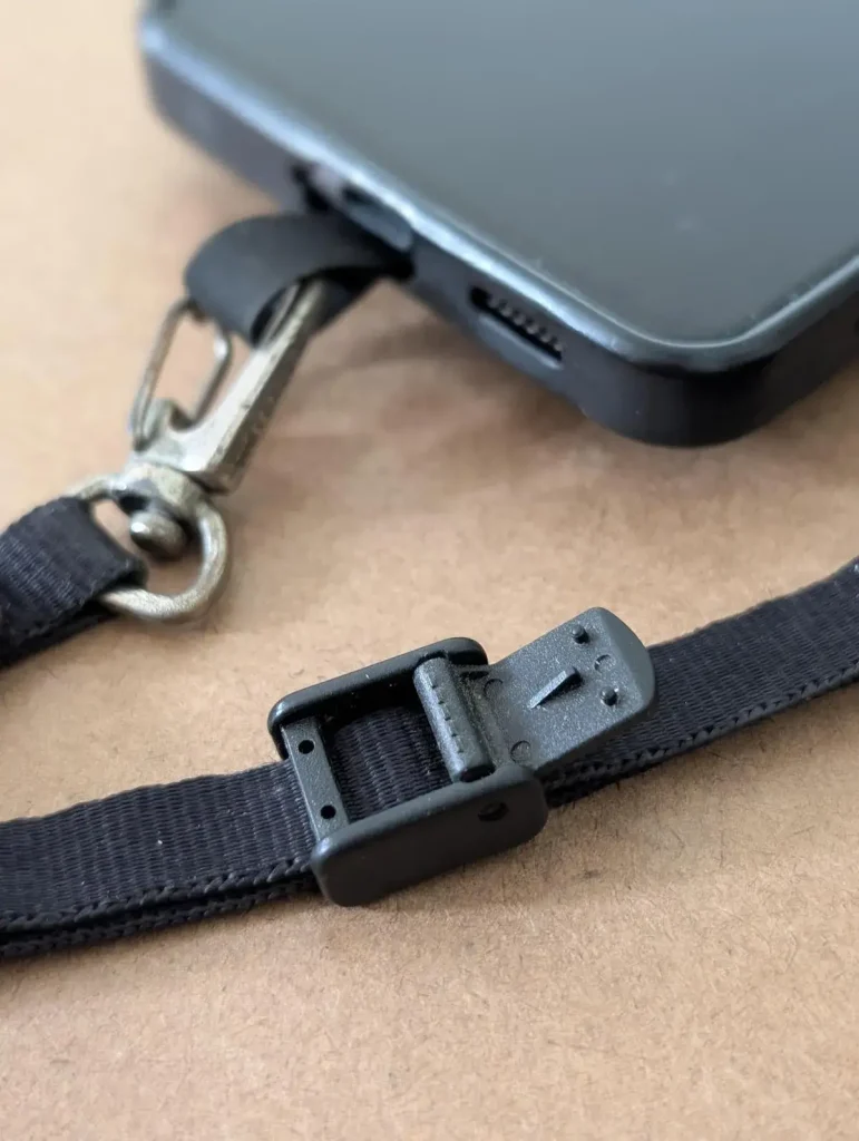A cellphone tether tab mounted to a cellphone with a small leash attached to it. The locking clip on the leash is open (in the not lock position where it can be adjusted by sliding it up or down the leash in a different position). This tether tab and leash combo is made by BlackRapid. The least has a quick-attach hook (similar to a rock climbing clip) on one end, and locking clip on the other end. You can slide your hand through the leash and use the locking clip to tighten it around your wrist, so that it stays on securely. Double-sided tape is provided. The tether tab is aligned with the bottom of the cellphone and meant to stick out where the charging port is. If a cellphone case is used, the tether tab will stick out through the hole for the charging port.