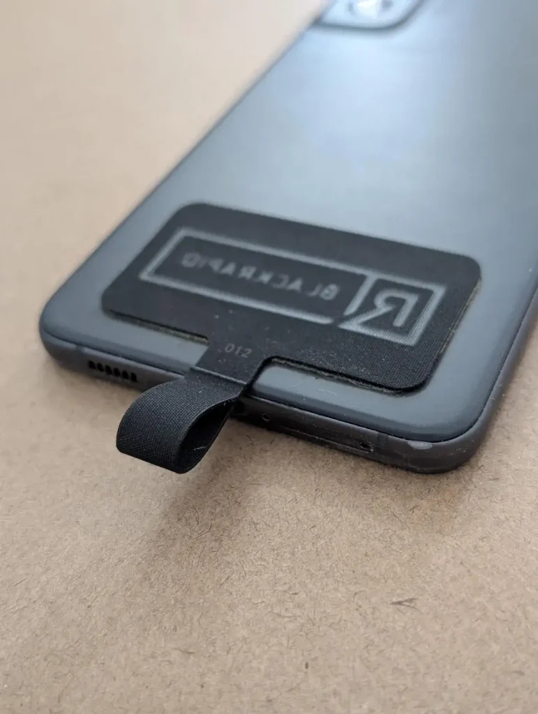 A cellphone tether tab mounted to a cellphone. This tether tab is made by BlackRapid. Double-sided tape is provided. The tether tab is aligned with the bottom of the cellphone and meant to stick out where the charging port is. If a cellphone case is used, the tether tab will stick out through the hole for the charging port.