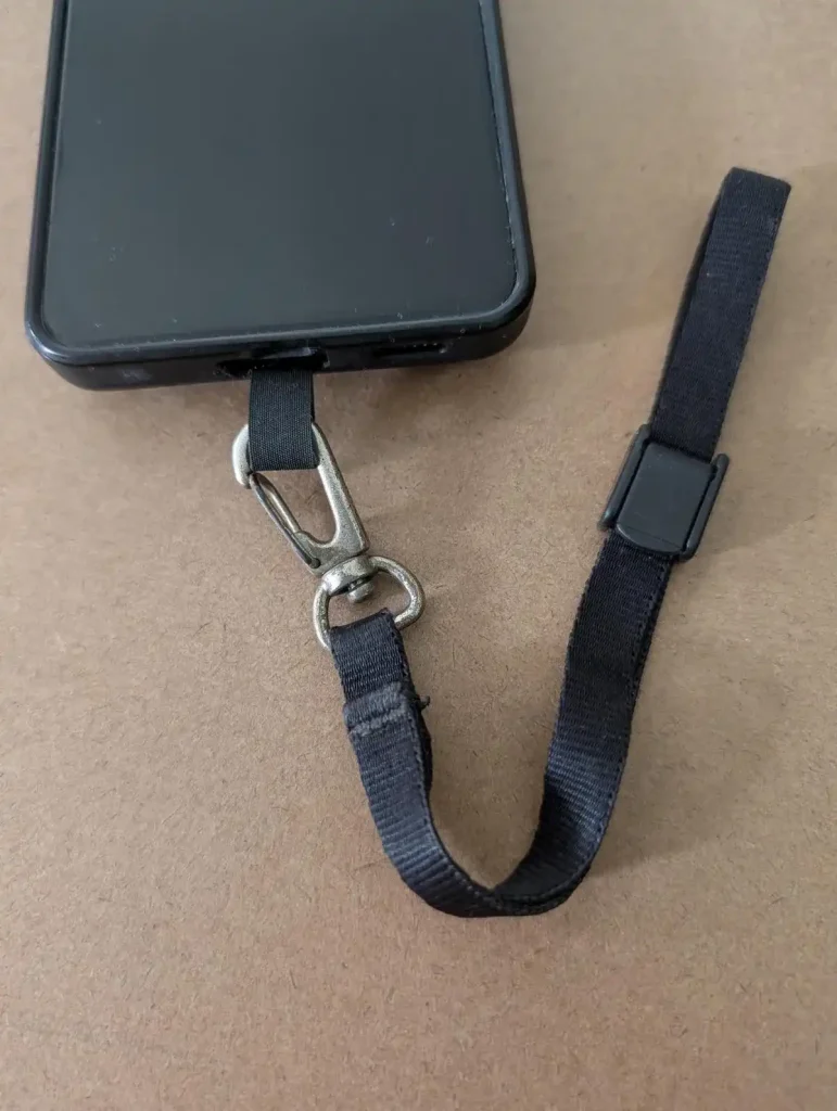 A cellphone tether tab mounted to a cellphone with a small leash attached to it. This tether tab and leash combo is made by BlackRapid. The least has a quick-attach hook (similar to a rock climbing clip) on one end, and locking clip on the other end. You can slide your hand through the leash and use the locking clip to tighten it around your wrist, so that it stays on securely. Double-sided tape is provided. The tether tab is aligned with the bottom of the cellphone and meant to stick out where the charging port is. If a cellphone case is used, the tether tab will stick out through the hole for the charging port.