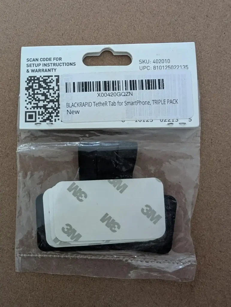 A pack of three BlackRapid-brand tether tabs with double-sided tape. This image is the backside of the pack, and shows the double-sided tab in front of the tabs. The package has a SKU number, 402010, UPC number 810125022135. There is a QR code that can be scanned for setup instructions and warranty.