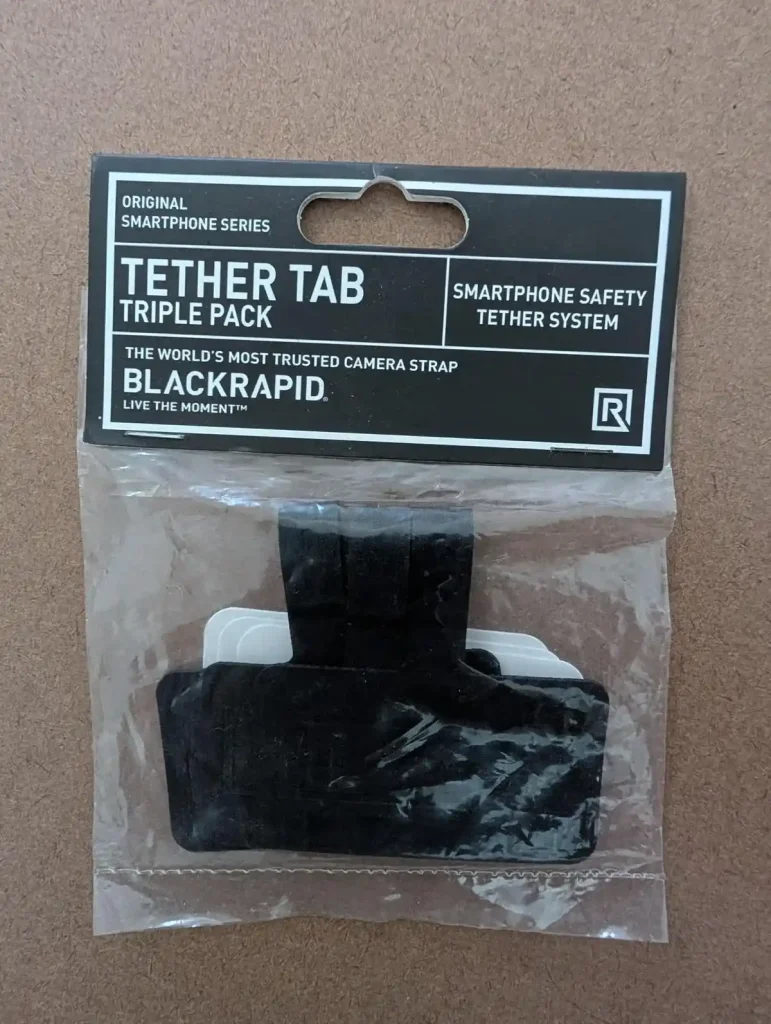 A pack of three BlackRapid-brand tether tabs with double-sided tape. This image is the front of the pack, and shows the tabs. The package reads: "Tether Tab Triple Pack", "Smartphone Safety Tether System", The Word's Most Trusted Camera Strap BlackRapid", "Live the moment".