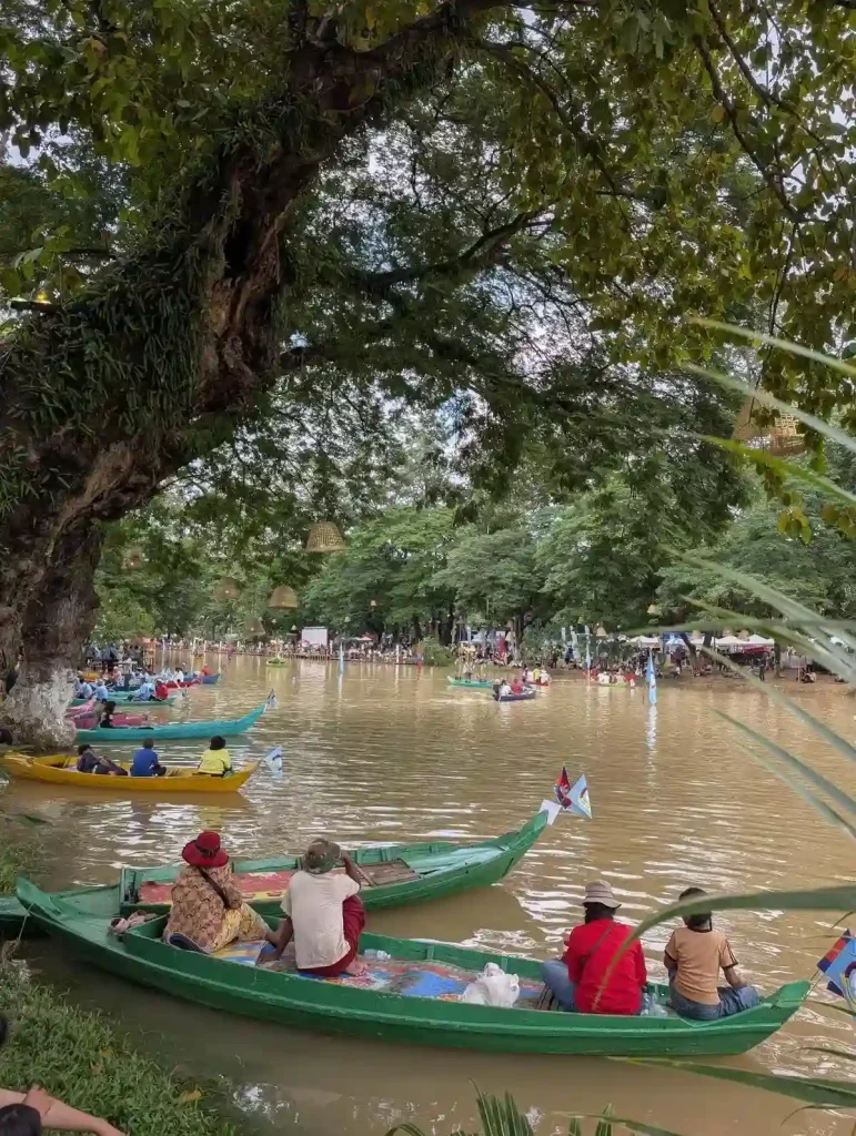 People sitting in boats along a shore watching a boat race. There is a massive tree that arches over into the river, which they are parked under. There are five boats that are spaced apart. This is along the the shore of the Siem Reap river during the Boat Racing festival (Bon Om Touk) in Siem Reap, Cambodia. There are no boats racing in this image.